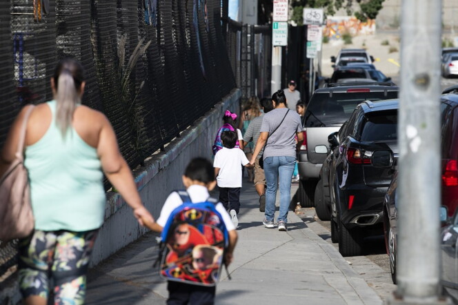 parents walk their children along a sidewalk with a chainlink fence on one side and a row of cars on the other side.