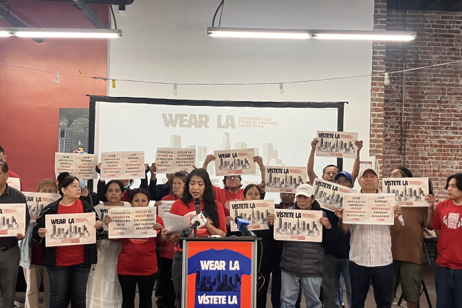 A group of people, some in red shirts, stand behind a podium holding signs that say "Wear LA" and "Visiete LA" with images of the Los Angeles skyline. One woman with a medium skin tone and long dark hair speaks at a microphone. There is a screen behind them that also says "Wear LA."
