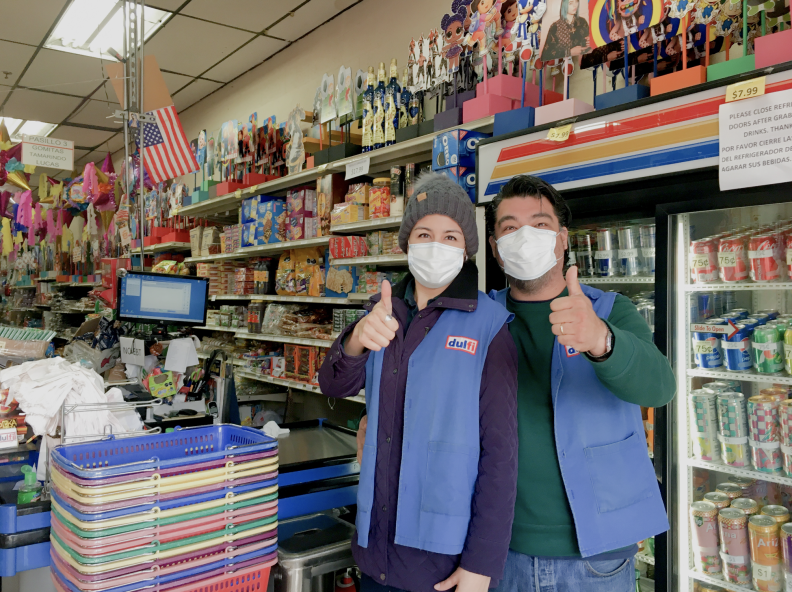 A man and woman in blue vests giving a thumbs up inside a store, by the cash register.