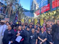 A crowd of people are standing before a large hotel in downtown Los Angeles. There are several signs and a flag as  well as palm trees lined behind them with a blue sky peaking behind them. The front row shows five women smiling. 
