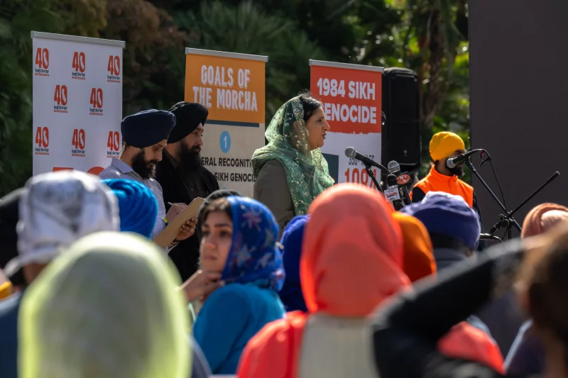 People in head scarves and turbans gather outside in a crowd. There are a couple people on stage behind the mics