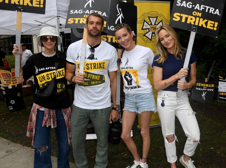 Four people with light-tone skin wear T-shirts that say SAG-AFTRA on strike on and carry picket signs