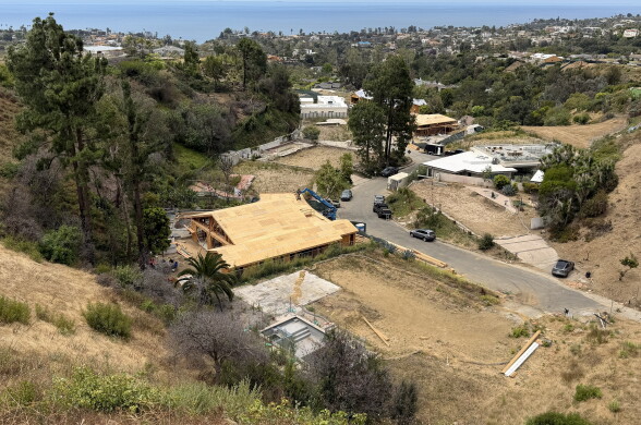 The wood frame of a home sits on a street with mostly empty lots surrounded by fencing. The ocean is visible in the distance.