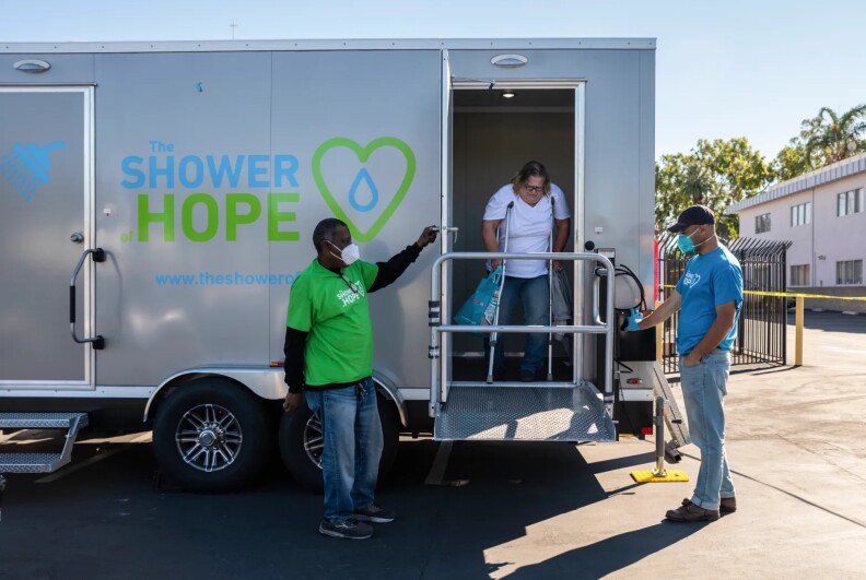 A woman on crutches comes out of a struck with text on it reading "THE SHOWER HOPE," an icon of a shower, an icon of a green heart with a giant water drop inside it, and a web address that begins "www.theshower" before being cut off by a Black man in a green shirt standing in front of it. To the right, a light brown-skinned man stands wearing a light blue T-shirt.