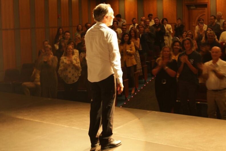 A man stands on stage in the spotlight, facing award from the camera. The audience he's addressing is giving him a standing ovation.