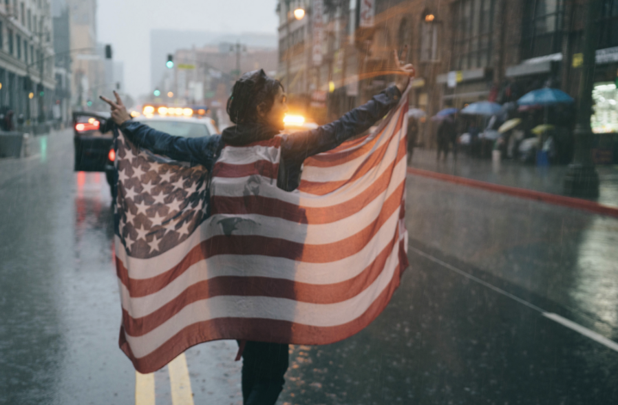 A woman stands in the middle of a road as it rains. She holds up an American flag and is raising both her hands in the peace sign. Behind her a police car can be seen parked and passersby with umbrellas. 