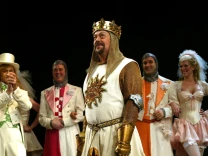 Tim Curry, wearing a costume of a king, looks up out of frame as other performers stand on stage behind him.