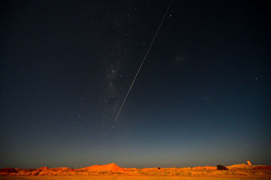 A bright white line streaks across a darkened sky over a barren, reddish-brown landscape.