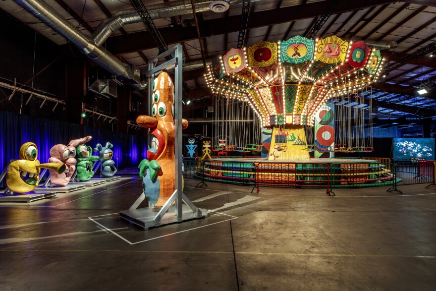 Various fantastical, cartoon faces with exaggerated facial features and varying numbers of eyes appear in front of a lit-up carousel in a warehouse space.