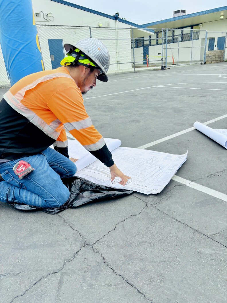 A person crouches on a blacktop and points to sheets of white paper.