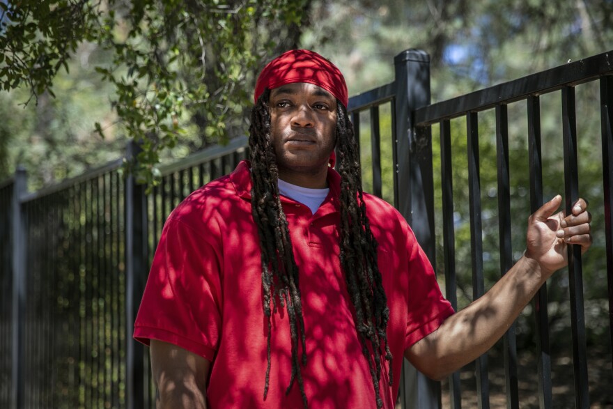 A Black man who appears to be in his 30s or 40s stands next to a wrought iron gate, loosely holding one of the bars. His hair is in long locs and he wears a red polo shirt and red hat. He gazes to the left of the photo and is unsmiling. It is daytime and lush green trees can be seen behind him.