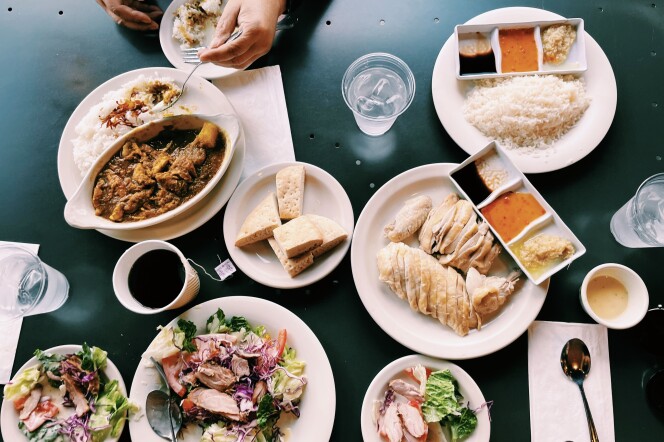A hand reaches out with a fork to dishes with rice, chicken and veggies in Savoy Kitchen. The dishes sit on a green table with utensils and sauce.