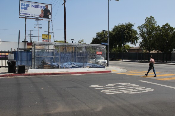 A woman walks in a crosswalk next to a fenced off entrance to a pedestrian tunnel. A blue tarp, bicycle and other items are visible through the fence.
