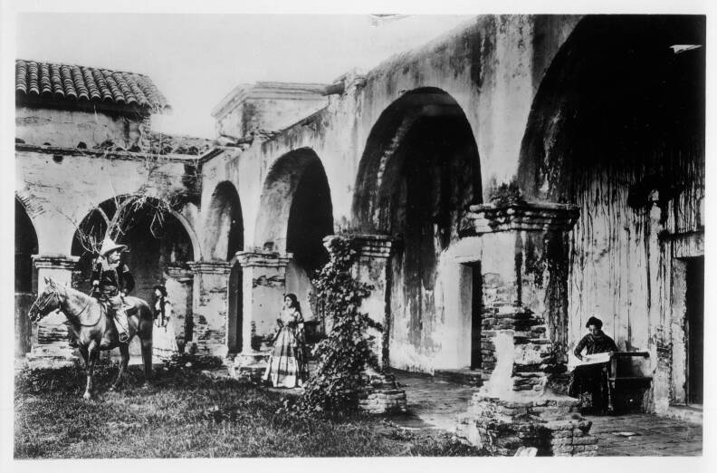 A man sits on a horse in the courtyard while two young women in long dresses stand under the arches of a large stone building. Another woman sits on a bench nearby.