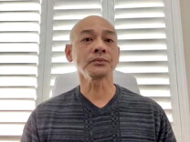 A 48-year-old man of Vietnamese descent with a shaven head looks somberly into the camera, with a window with slatted blinds behind him. 