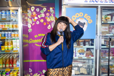 An Asian woman with dark brown hair stands in front of refrigerator doors inside a grocery store, holding a microphone, wearing a blue sports jacket. In the background are coolers filled with frozen foods, sodas, and a sign that reads El Sereno Green Grocer.