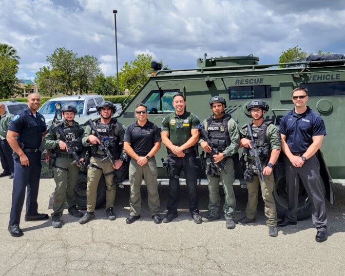 A row of men, most holding weapons, post in front of an armored vehicle.