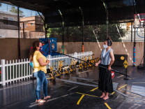 a woman in a yellow shirt and jeans talks into a microphone held on a long boom pole by a woman in a black skirt and striped shirt standing 6 feet away. both of them wear face masks