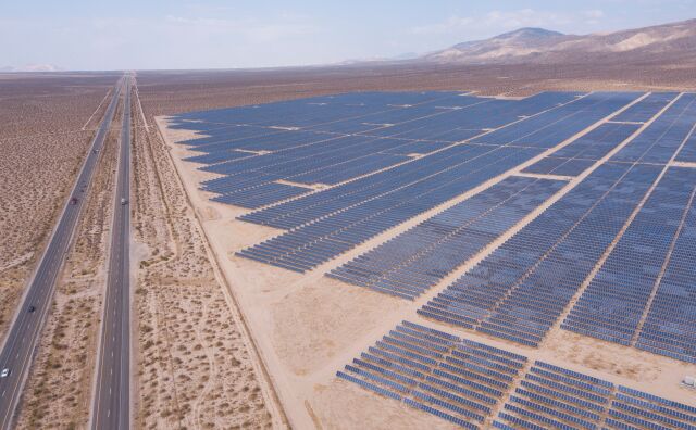 A field of solar panels is surrounded by dry flat fields with mountains in the background.