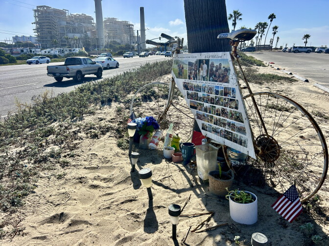 A white, rusted bicycle frame decorated with photos and 'A Life Well Lived' sign stands as a roadside memorial with flowers, flags, and candles.