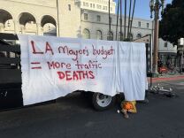 A man with an orange helmet lays on the road behind a truck with a white rose in his right hand. The truck is draped with a white sheet and the following painted on it in red: LA Mayor's budget = more traffic DEATHS.