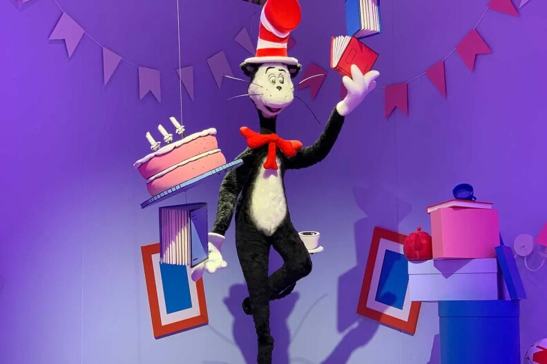 A giant puppet version of the Cat in the Hat character from Dr. Seuss books stands on a ball and holds a book in hand in a room full of streamers, boxes, with picture frames on the walls behind him. He is black with a white belly and is wearing a red and white-striped top hat.