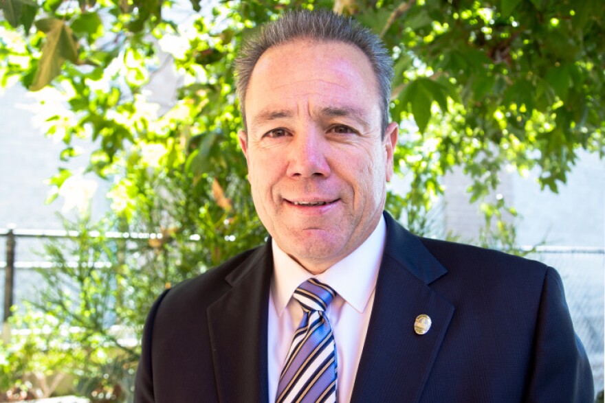 A middle-aged white man with close cropped graying hair wears a blue suit with a pin on the lapel