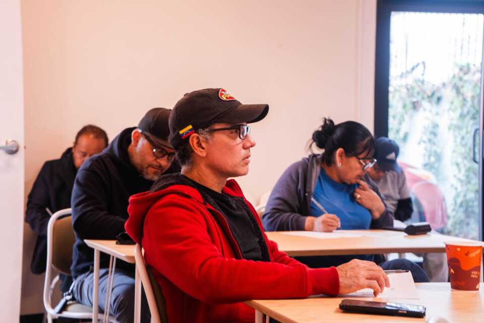 A person sits at a desk with others around him. The person wears a baseball cap and a red sweatshirt.