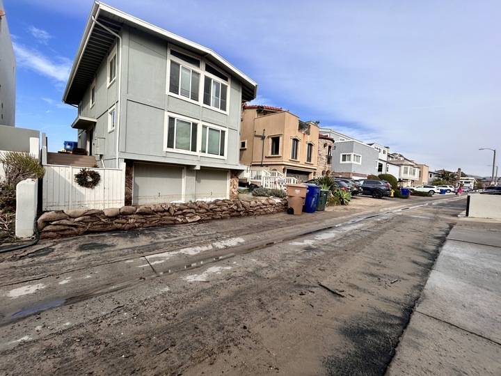 Sandbags line the front of a grey home in a row of houses. Two garbage cans sit on the street in front of the home and a Christmas wreath hangs on the home's white fence.