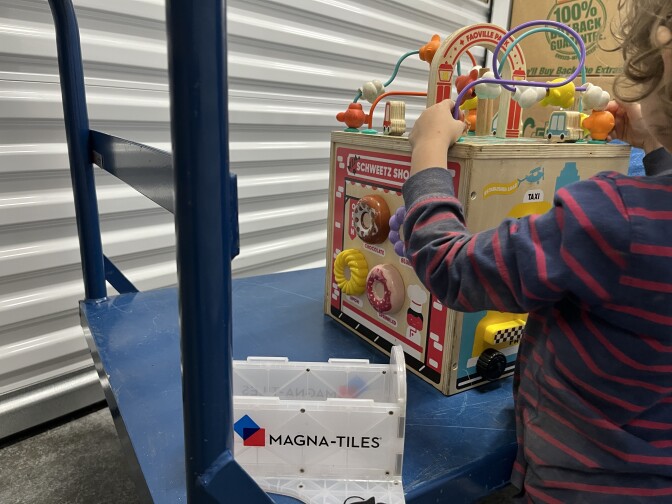 A young child with a dark blue and red striped shirt plays with wooden activity toy atop a blue steel dolly.