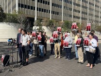 Two people stand behind a portable mic stand, one is clad in a suit and tie, the other has lifted their pant leg to reveal an ankle monitor. Behind them, about a dozen people hold up red, black, and white signs that read: "Drop All Charges Against John"