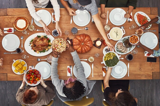 An overview of a dining table with people passing plates to one another. On the table, along with white dinner plate settings, is the typical Thanksgiving feast - a turkey, corn, pumpkin pie, ham, carrots, mashed potatoes and a large pumpkin in the middle.