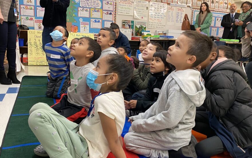 A group of about a dozen young children in first grade are seated on the floor of a classroom looking up. Two children are wearing blue masks. Several adults stand along the walls, which are covered with posters.
