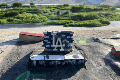 A picture of a miniature skate park is seen with a relatively large "LA" sign in Dodger Blue in the center. A brick obstacle can be seen to its left and a brick ramp can be seen to its right. The LA River is seen in the background, there's a lot of greenery growing out of the center of the river. The other side of the river can be seen sloping upwards.