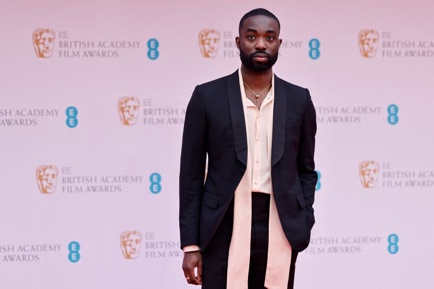 A man with dark skin tone stands in front of a pinkish backdrop that features the words "British Academy Film Awards."