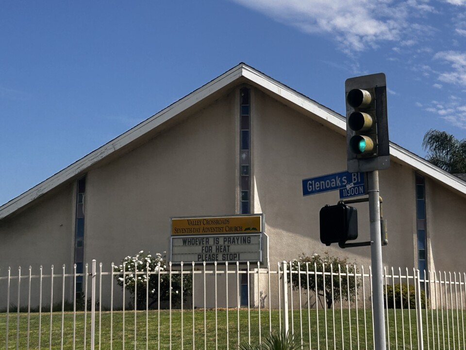 An image of an A-framed church with a green lawn and white metal fence on a sunny day with blue skies. A sign in front of the church reads "Whoever is praying for heat please stop!"