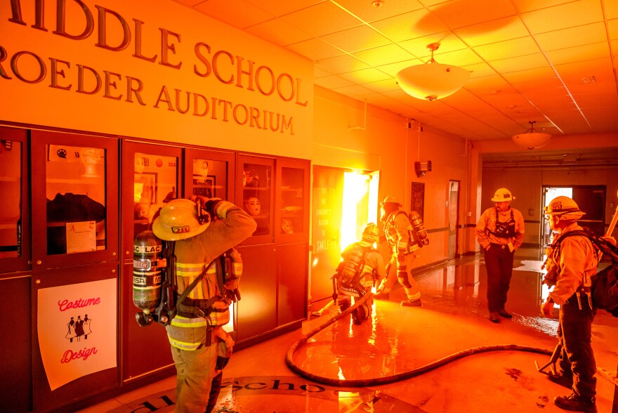 Fire fighter stand in a school hallway and open a door filled with fire inside. The wall in the hallway reads "Middle School/ Auditorium."