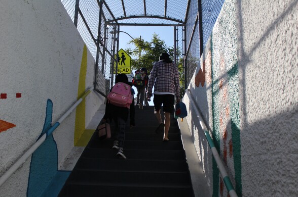 Two children wearing backpacks and an adult holding a small backpack walk up the stairs from an underground tunnel, with colorful art on the walls.