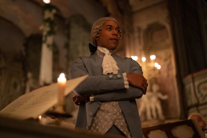 Kelvin Harrison Jr. stars in the film CHEVALIER. In this picture, he stands with his arms crossed in a candlelit room in 18th  century costume and wig.  