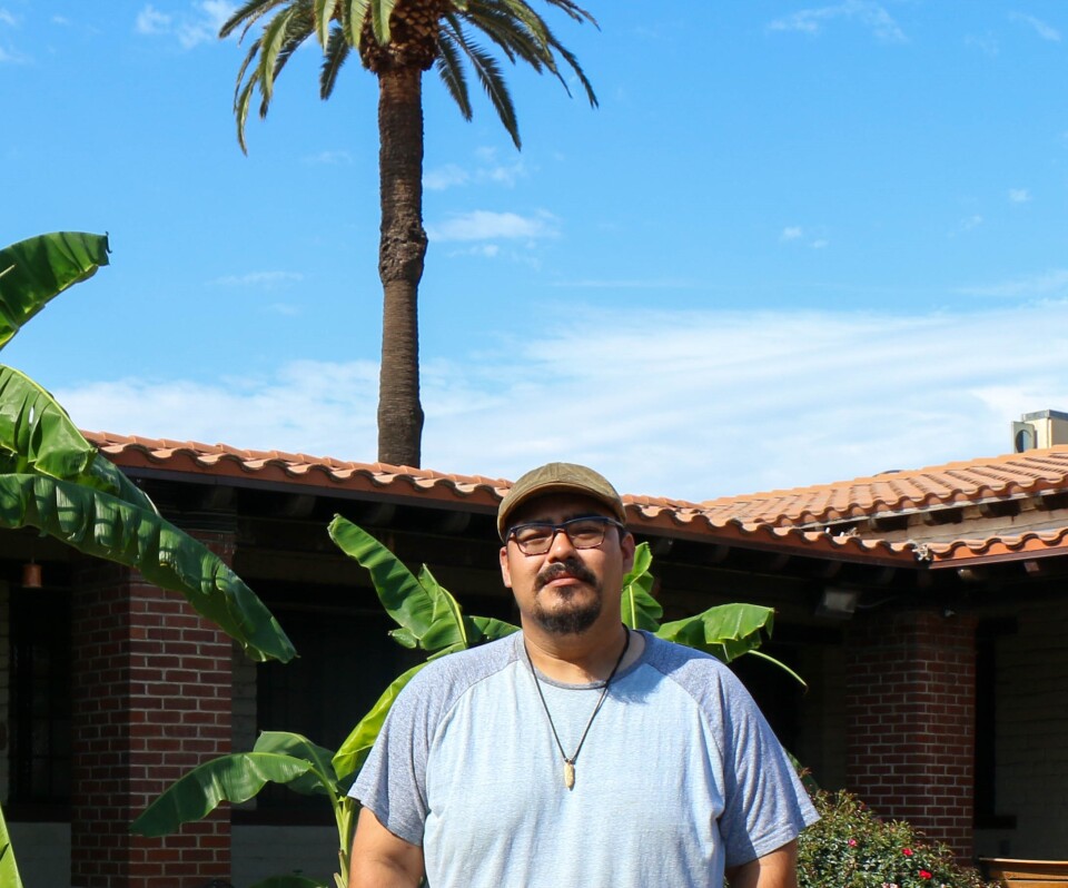 A man with light brown skin, black-rimmed glasses and a tan cap wears a light blue t-shirt and necklace under a sunny blue sky. Adobe and brickk building in the background with palm trees. 