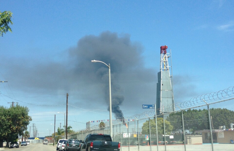 A oil drilling rig looms as smoke from a flare burns in the background of a residential street.
