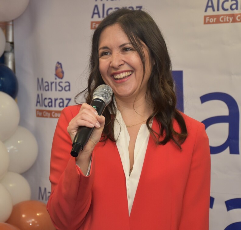 Marisa Alcaraz, a light-skinned Latina with brown hair falling just below her shoulders, smiles as she holds a microphone in front of a large banner with her name on it. Blue, white and orange balloons are behind her off to the side. She is wearing an orange coat over a white blouse.
