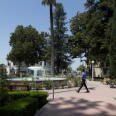 A man wearing black pants and a blue long sleeve walks across a plaza with a large water fountain at its center. 