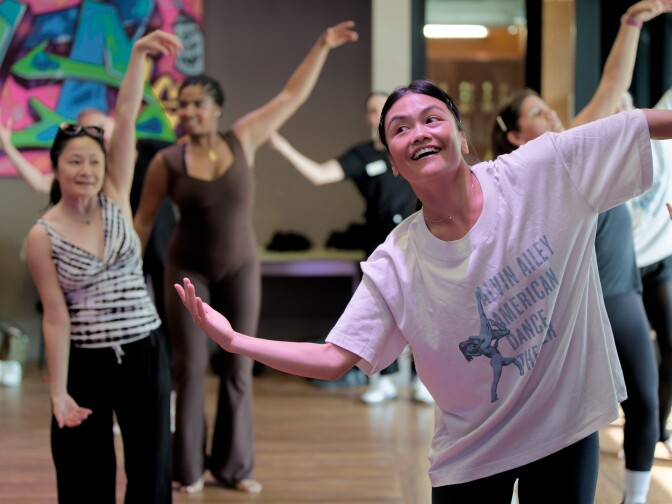 A smiling woman wearing an Alvin Ailey American Dance Theater t-shirt leads a group of people in a dance or movement class. They are all indoors with their arms raised and extended in various poses.