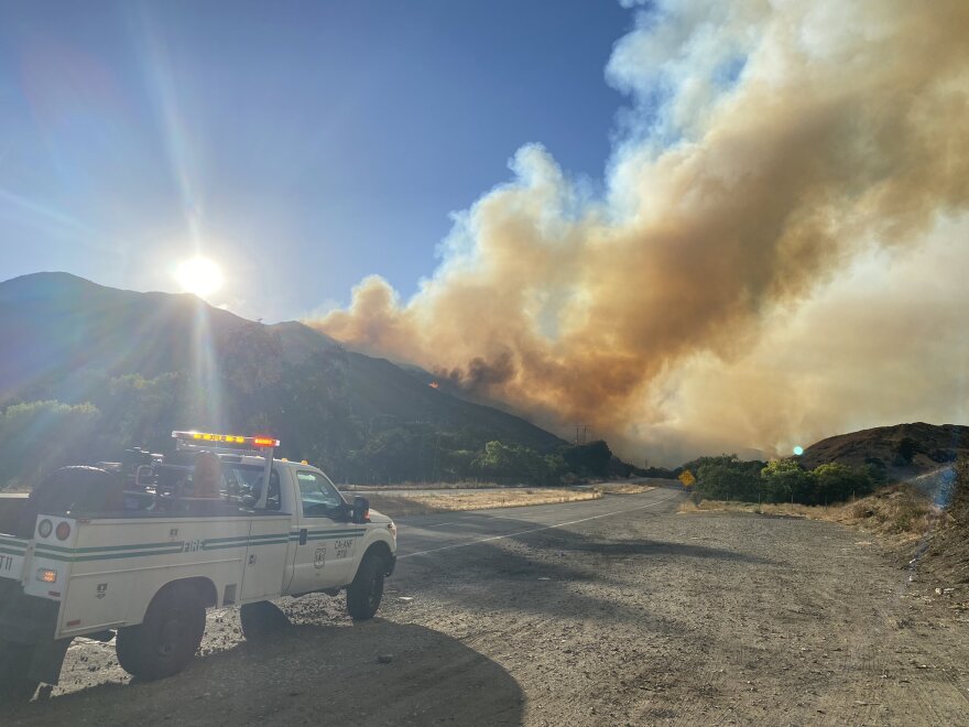 Smoke from a wildfire rises from a mountainside in the distance with a U.S. Forest Service truck parked in the foreground.