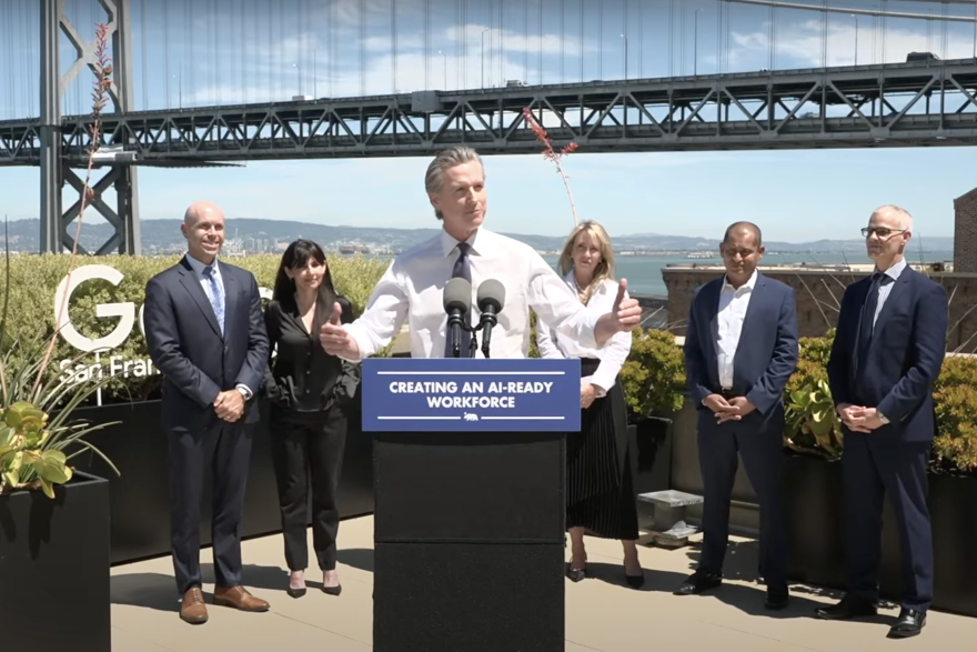 A man with light skin tone, clad in a white dress shirt and tie, gesticulates while speaking from a podium. On it, a label reads: "CREATING AN AI-READY WOKRFORCE." Five people stand behind him, also in business attire. The ocean and a bridge can be seen in the background.