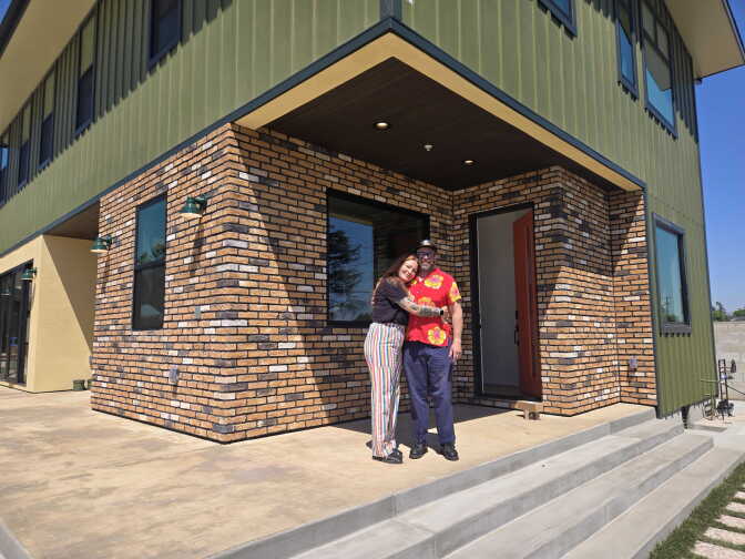 A man and a woman pose for a photo embracing each other standing in front of a two-story home with a brick exterior by the front door.