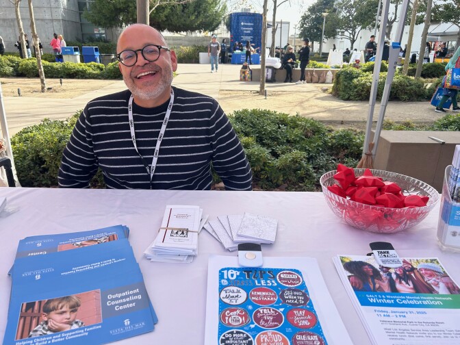 A man with medium light-skin tone and  black-framed reading glasses smiles while sitting behind a table. On it, there is a bowl with chubby red stars for squishing, along with handouts that read "Outpatient Counseling Center" and "10 Tips to Stress Less."