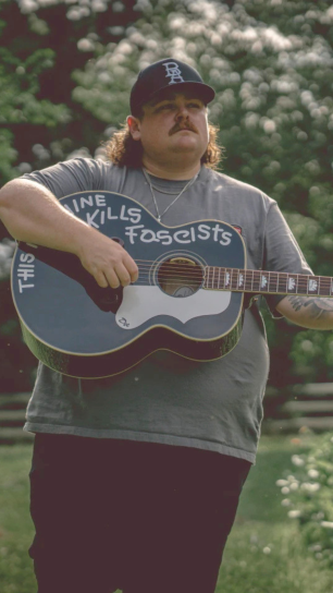A man with a mustache and mullet, wearing a ball cap and gray T-shirt, stands outdoors playing an acoustic guitar with "This machine kills fascists" hand-painted on the body, greenery and white blossoms in the background.