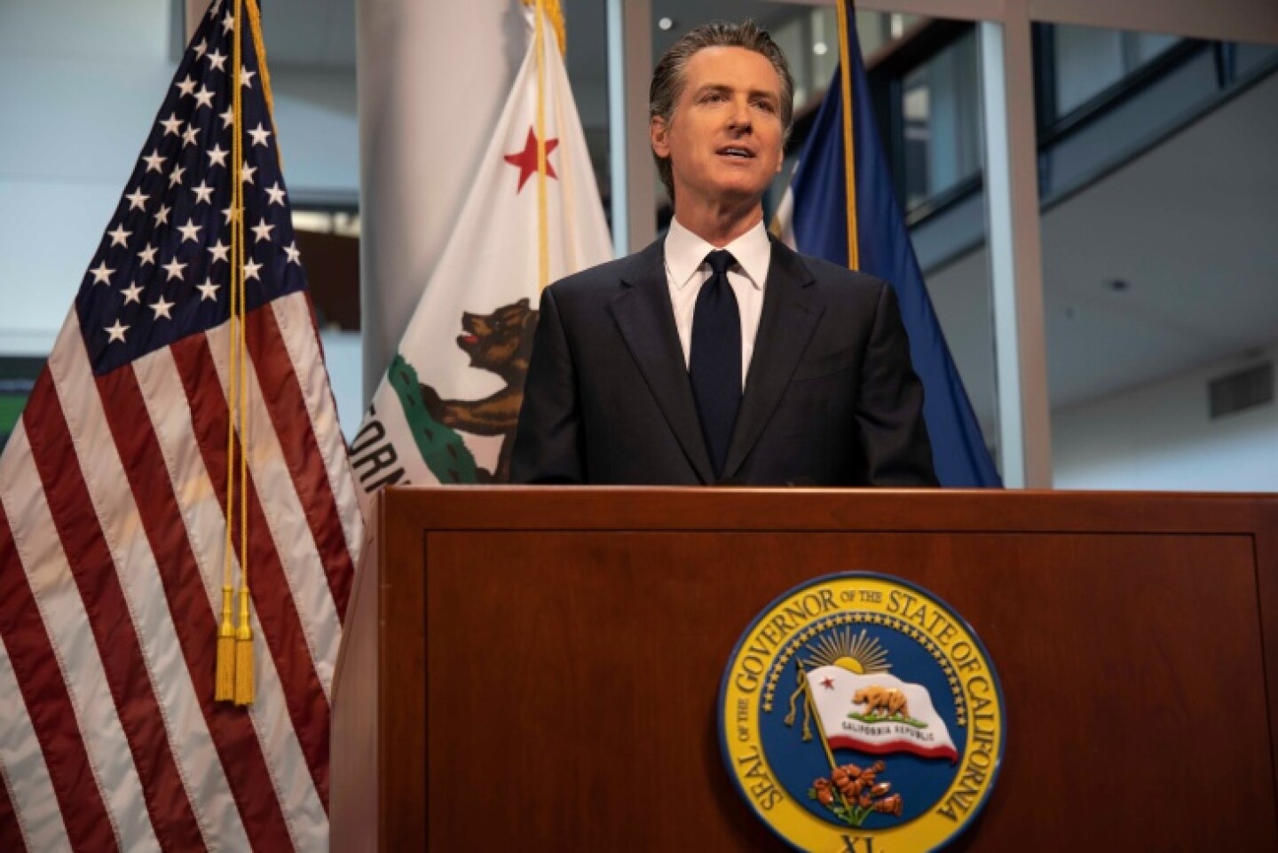 Gavin Newsom stands at a podium adorned with the seal of the governor of California, with the state and U.S. flags behind him